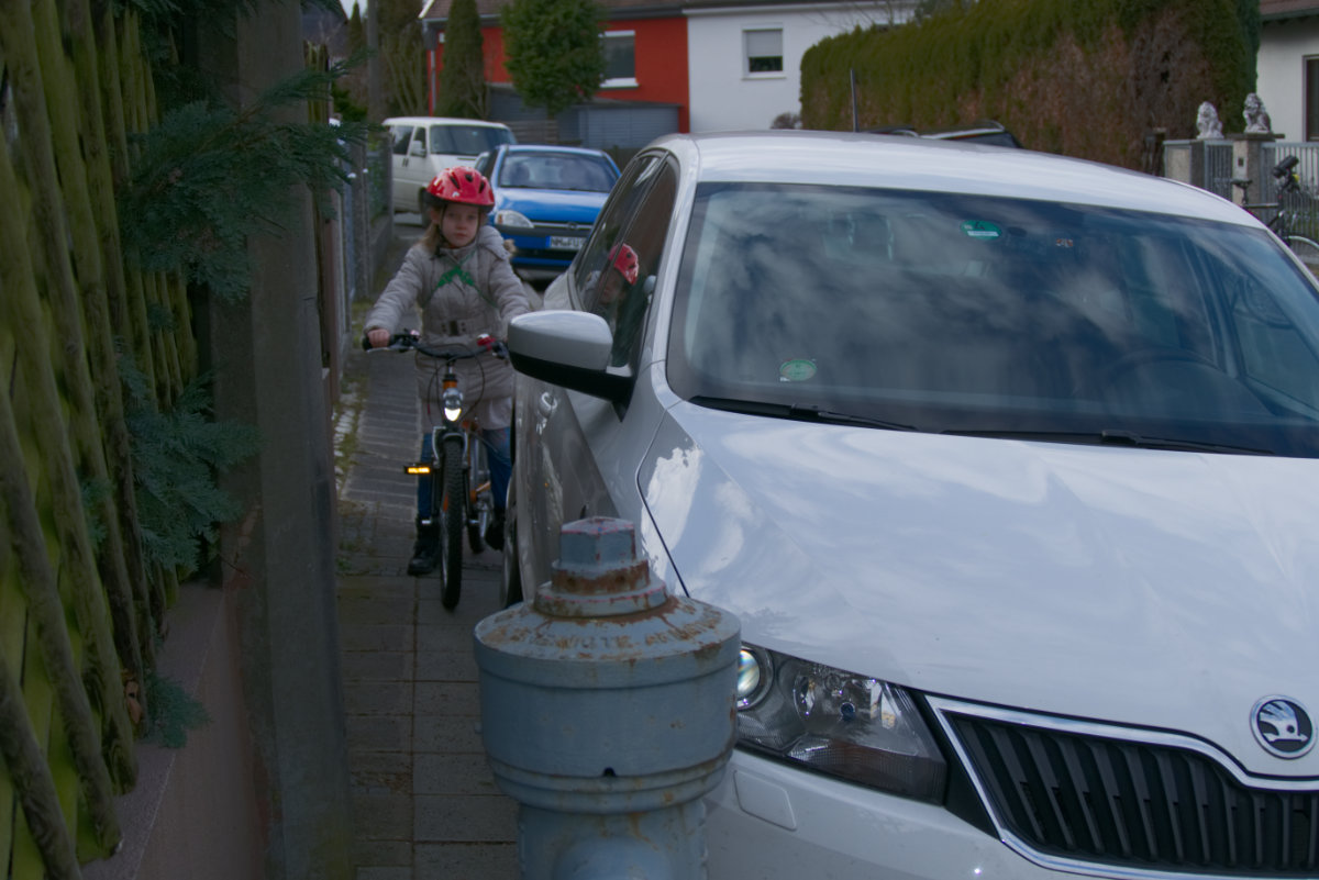 Foto eines Autos, das auf dem Gehweg parkt und so das Rad fahrende Kind am weiterfahren hindert.